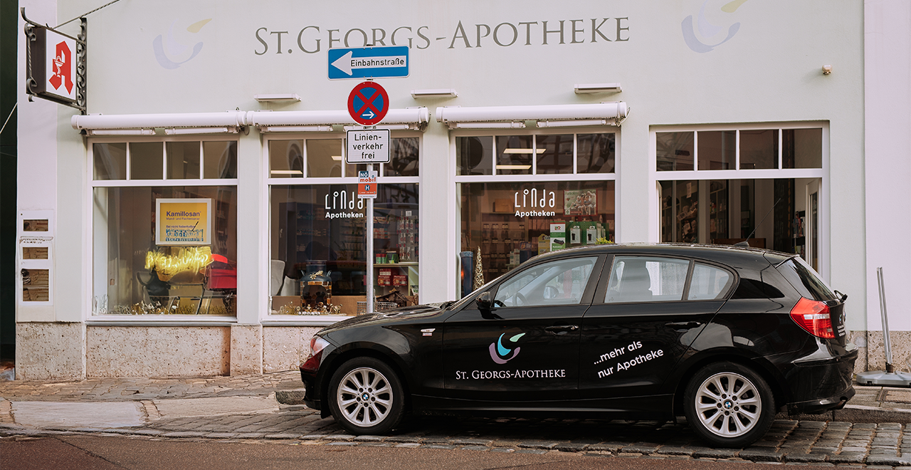 Außenansicht der St.-Georgs-Apotheke mit großen Schaufenstern. Vor dem Gebäude steht ein schwarzes Auto mit Apothekenlogo und der Aufschrift „mehr als nur Apotheke“.
