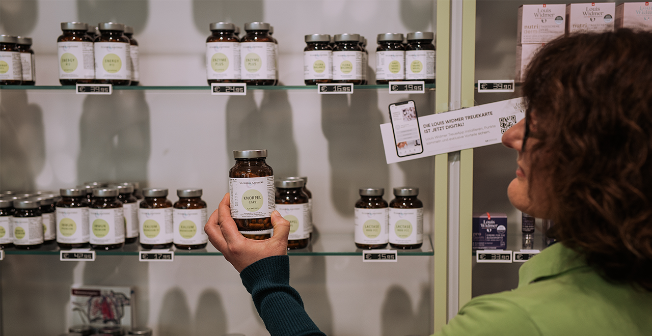 Eine Person steht vor einem Regal in einer Apotheke und hält ein braunes Glas mit Kapseln in der Hand. Im Hintergrund sind mehrere Reihen mit Nahrungsergänzungsmitteln in ähnlichen Gläsern zu sehen.