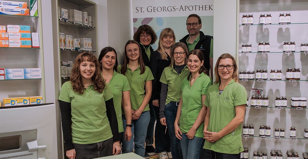Eine Gruppe von zehn erwachsenen Personen steht dicht beieinander in einem hellen Apothekenraum. Die meisten tragen grüne Arbeitsoberteile, einige schwarze Westen. Hinter ihnen sind Regale mit Medikamenten und Pflegeprodukten sowie ein Schild mit der Aufschrift „St. Georgs-Apotheke“.