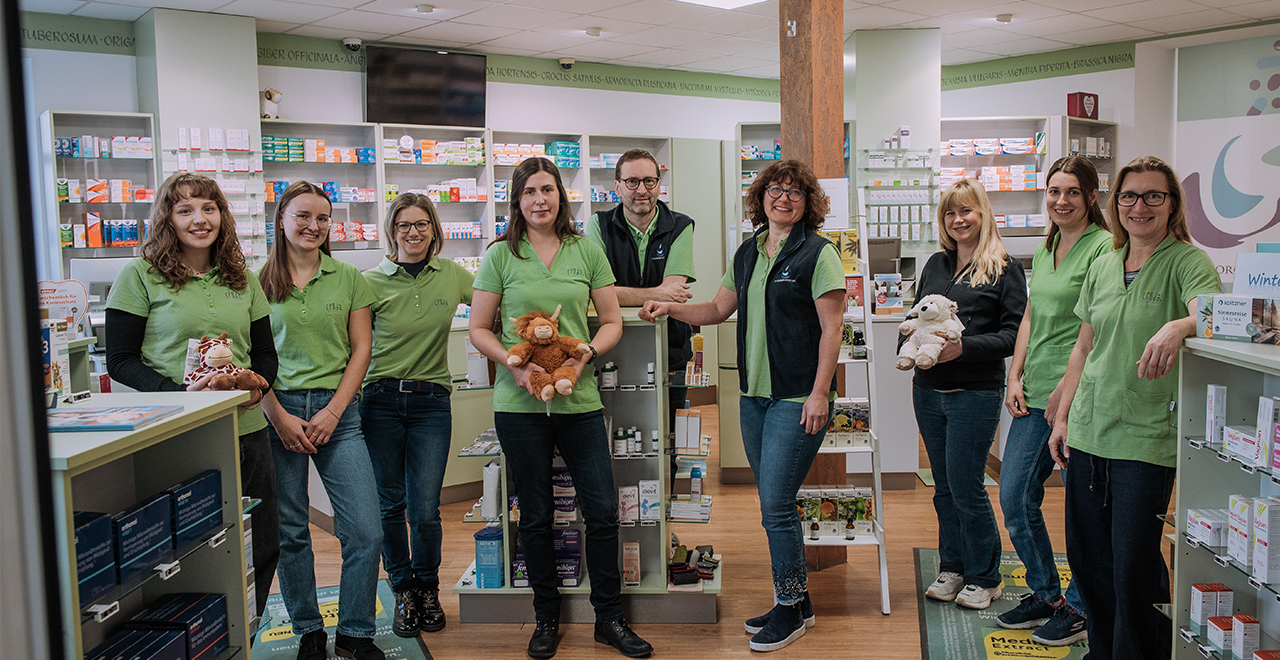 Neun erwachsene Personen stehen verteilt in einem Verkaufsraum einer Apotheke zwischen Regalen mit Arzneimitteln. Mehrere tragen grüne Arbeitskleidung, einige schwarze Westen. Zwei Personen halten Stofftiere in den Händen. Der Raum ist hell beleuchtet und übersichtlich eingerichtet.