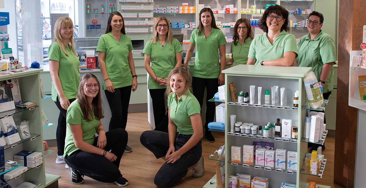 Neun Mitarbeiterinnen und Mitarbeiter in grüner Arbeitskleidung stehen lächelnd im Verkaufsraum einer Apotheke vor Regalen mit Medikamenten.
