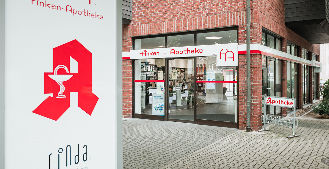 Eingangsbereich der Finken-Apotheke mit großer Glasfront, automatischer Tür und gut sichtbarem roten Apothekenzeichen auf einem weißen Schild im Vordergrund. Die Apotheke gehört zur LINDA-Apotheken-Gruppe.
