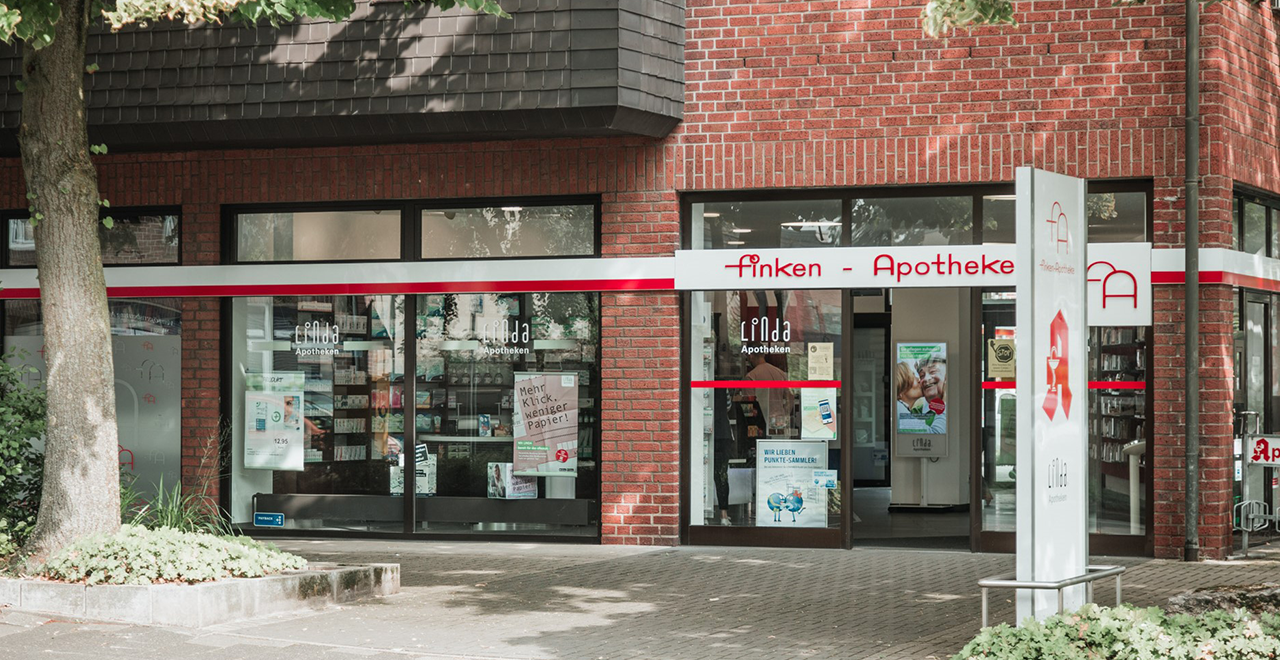 Frontalansicht der Finken-Apotheke mit Glasfassade und roten Akzenten auf weißem Hintergrund. Ein großes Schild mit Apothekenlogo steht im Eingangsbereich, daneben ein Fahrradständer mit dem Wort 