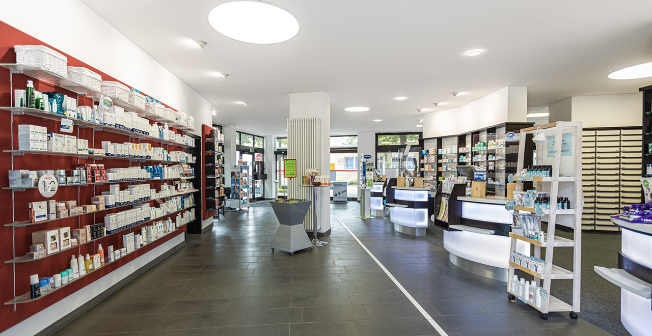 Helle Innenaufnahme der Apotheke mit Regalen voller Pflege- und Gesundheitsprodukte auf der linken Seite. Rechts befinden sich mehrere beleuchtete Beratungstheken. Im Hintergrund ist der Eingangsbereich mit großen Fenstern sichtbar.