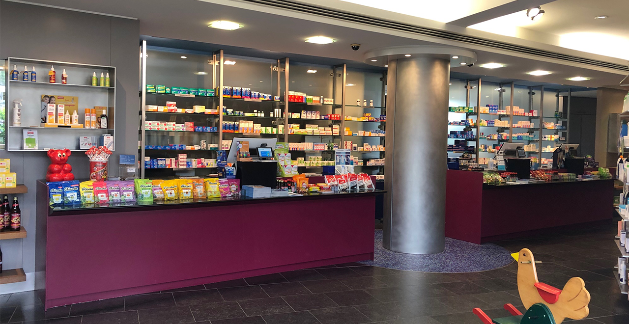 Verkaufsbereich einer modernen Apotheke mit Regalen voller Medikamente, einem langen Verkaufstresen und einem Spielzeug-Wipptier im Vordergrund.