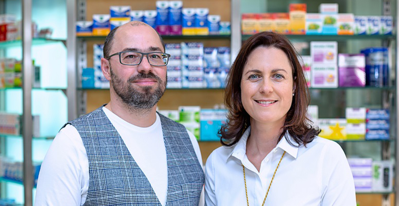 Der Inhaber der Apotheke steht zusammen mit seiner Frau in der Apotheke vor Regalen mit verschiedenen Medikamenten. Beide lächeln freundlich in die Kamera. Der Inhaber trägt eine graue Weste und Brille, während seine Frau ein weißes Hemd und eine goldene Halskette trägt. Die Szene vermittelt eine familiäre und professionelle Atmosphäre in der Apotheke.