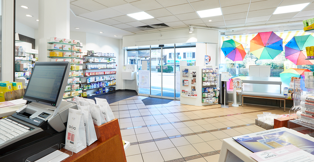 Innenansicht der Apotheke Rohr. Der Verkaufsraum ist gut beleuchtet und bietet Regale mit einer Vielzahl von Medikamenten und Gesundheitsprodukten. Ein Computerbildschirm ist am Verkaufstresen zu sehen, und bunte Regenschirme hängen im Fenster und verleihen dem Raum eine freundliche, einladende Atmosphäre.