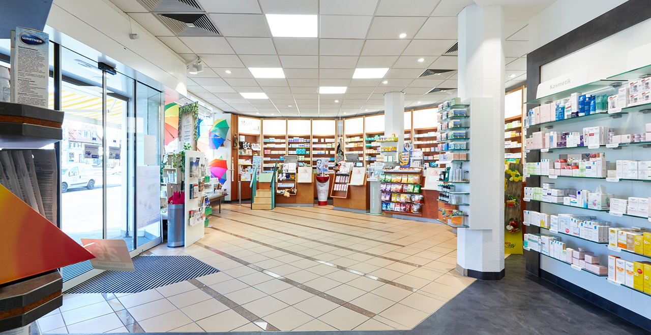 Innenansicht der Apotheke Rohr mit Regalen, die Kosmetik- und Gesundheitsprodukte an den Wänden präsentieren. Im Vordergrund stehen Ständer mit Artikeln für die tägliche Pflege, und das Design ist modern und aufgeräumt. Die Regale bieten eine große Auswahl an Produkten, die ordentlich und übersichtlich angeordnet sind.