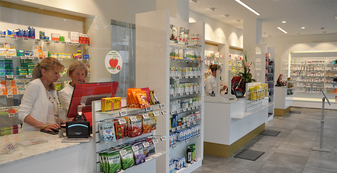 Zwei Apothekenmitarbeiterinnen stehen an einem modernen Verkaufstresen und bedienen ein rotes Kassensystem. Im Vordergrund sind Bonbons und Erkältungsprodukte in Regalen sichtbar. Rechts und im Hintergrund befinden sich weitere Verkaufsbereiche mit Regalen und einer dritten Mitarbeiterin, die ebenfalls Kundendienst leistet. Der Raum ist hell, ordentlich und barrierefrei zugänglich.