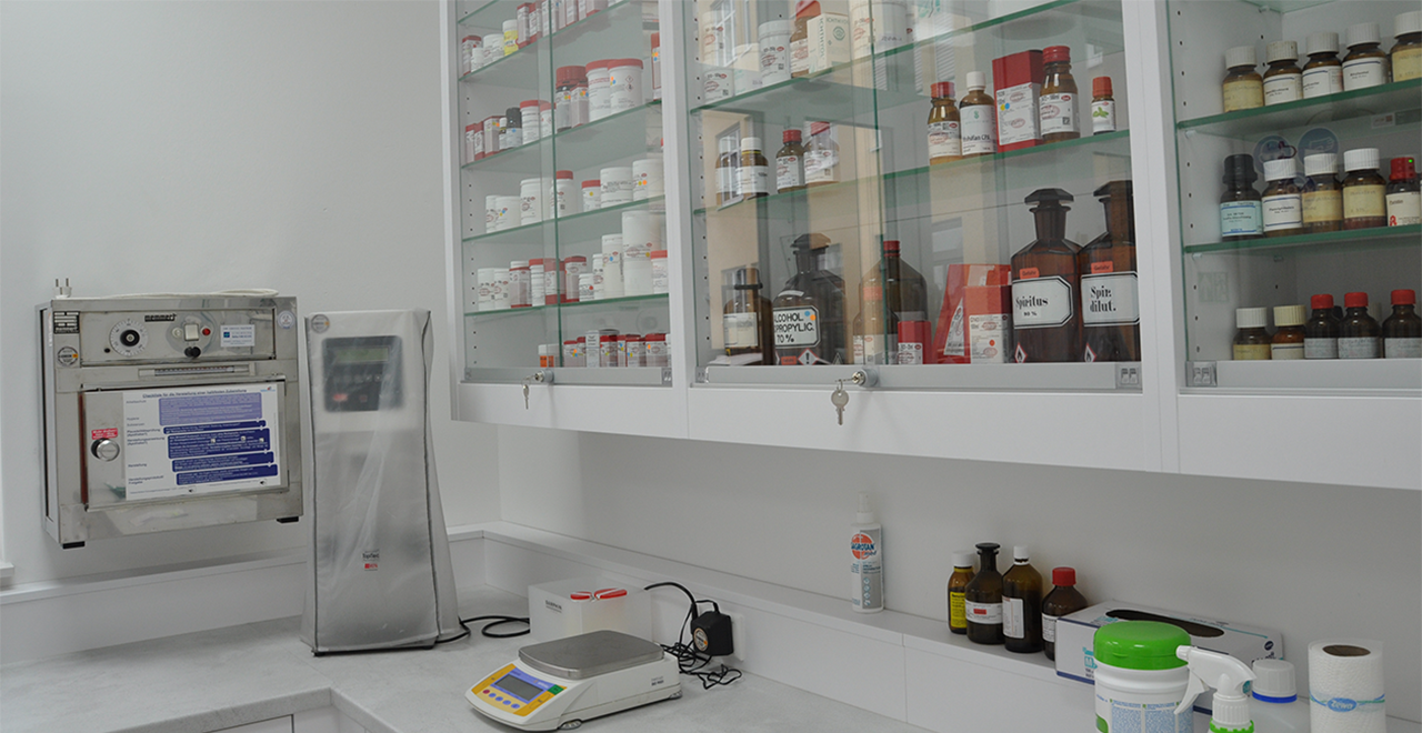 Blick in den Laborbereich der Apotheke mit verschiedenen Laborgeräten wie einer Waage, Mischgeräten und Glasregalen mit Rezeptursubstanzen. Die Umgebung ist hygienisch und sachlich eingerichtet, typisch für pharmazeutische Herstellung.
