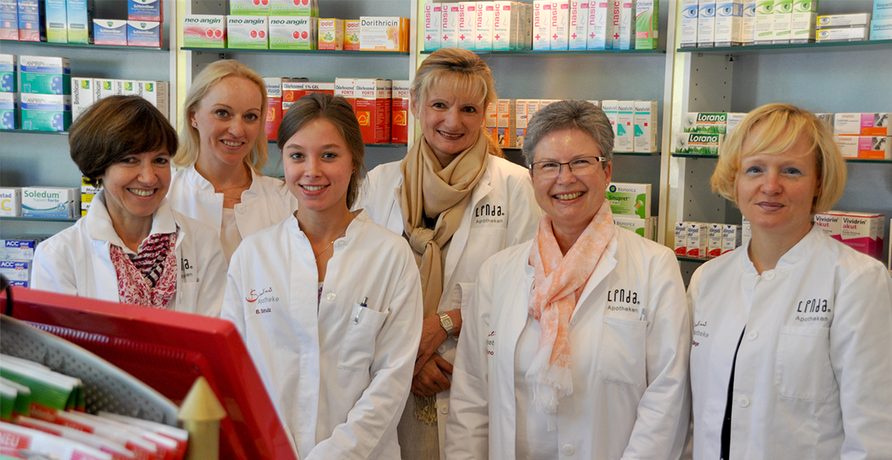 Sechs weibliche Apothekenmitarbeiterinnen in weißen Kitteln stehen lächelnd vor einem Regal mit Medikamenten. Auf den Kitteln sind Apothekenlogos und Namensschilder sichtbar. Die Stimmung ist freundlich und professionell.
