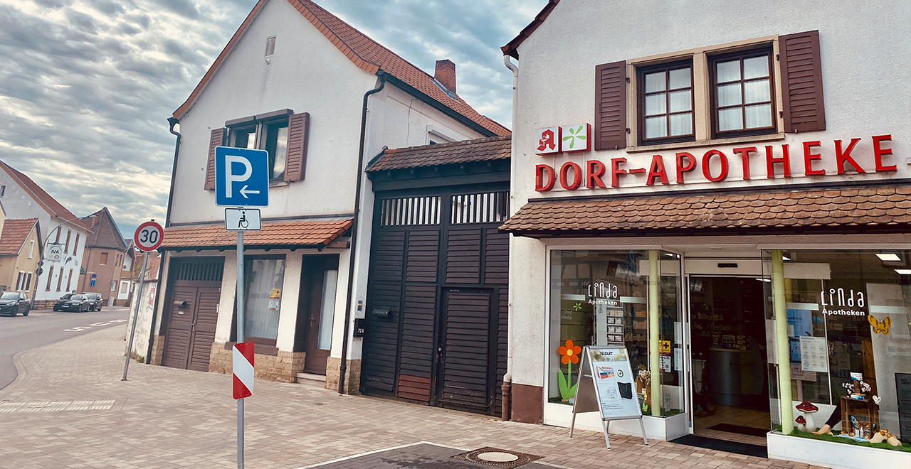 Eingang der Dorf-Apotheke in einem Wohnhaus mit rotem Ziegeldach. Der Schriftzug „Dorf-Apotheke“ ist in roten Buchstaben über der Tür angebracht. Ein Schild zeigt einen Behindertenparkplatz in der Nähe. Die Eingangstür ist barrierefrei.
