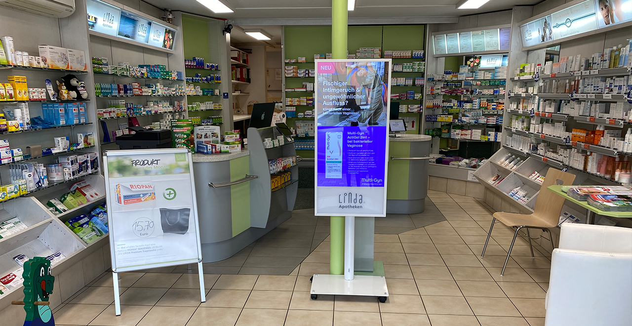 Innenraum der Dorf-Apotheke mit Verkaufsregalen voller Medikamente und Pflegeprodukte. In der Mitte steht ein digitaler Aufsteller mit Gesundheitsinformationen. Links steht ein Aufsteller mit dem „Produkt des Monats“. Der Verkaufsbereich hat eine geschwungene Theke.