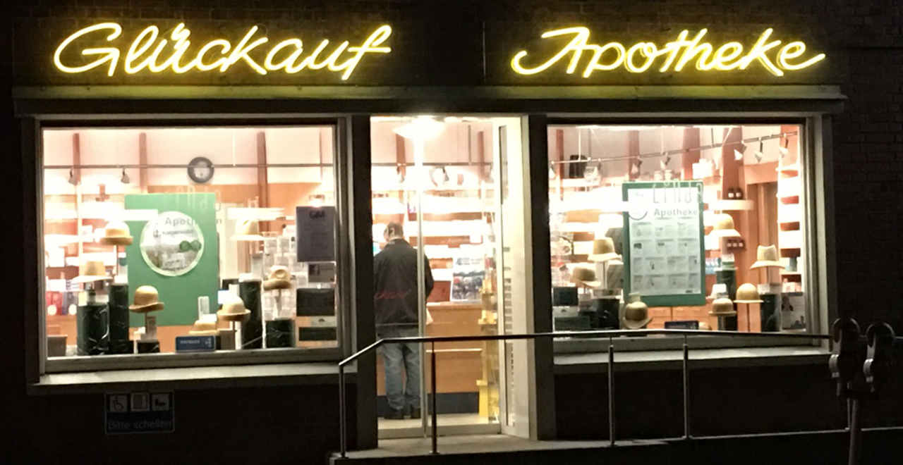 Die gleiche Apotheke „Glückauf Apotheke“ bei Nacht. Die gelbe Leuchtschrift über den Fenstern ist deutlich sichtbar. Die Apotheke ist innen hell erleuchtet, sodass viele Regale mit Medikamenten sichtbar sind. In der Mitte des Bildes steht ein Mann im Eingang der Apotheke, von hinten zu sehen. Im linken Fenster sind erneut dekorative Hüte sowie Werbematerial zu erkennen. Auch hier ist ein Geländer mit Rampe und das blaue Hinweisschild „Bitte schellen“ zu sehen.
