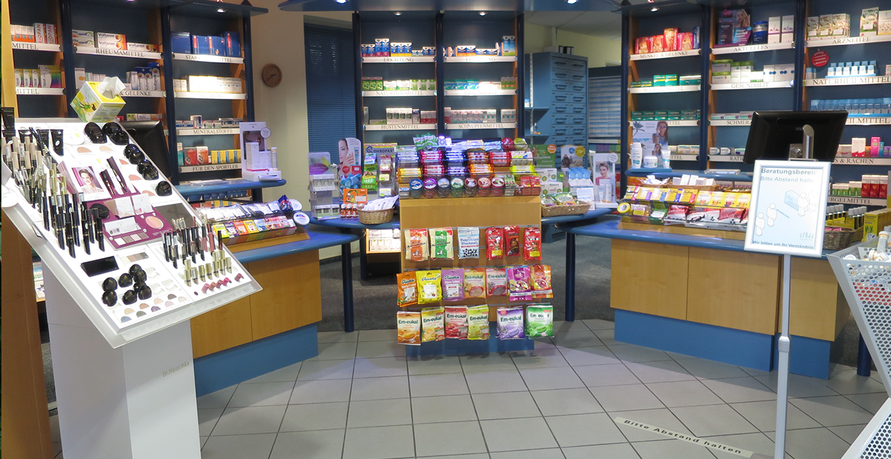 Innenraum einer Apotheke mit Verkaufsregalen, Kosmetikaufsteller und Hinweisschild zum Beratungsbereich.
