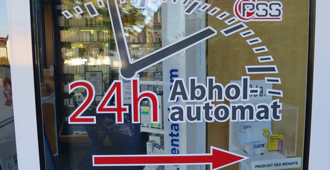 Schaufenster mit Aufschrift ‚24h Abholautomat‘ und Blick auf Medikamente im Hintergrund.
