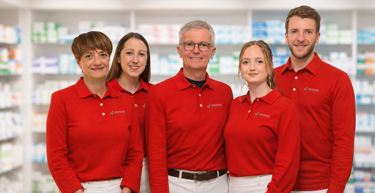 Fünf Mitarbeitende einer Apotheke stehen nebeneinander vor Regalen mit Medikamenten. Sie tragen rote Poloshirts mit Apothekenlogo und weiße Hosen und lächeln in die Kamera. Im Hintergrund sind unscharf gefüllte Apothekenregale zu sehen.