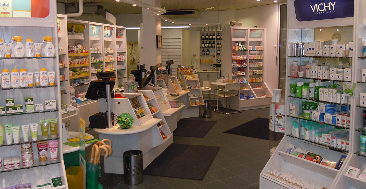 Innenansicht einer Apotheke mit verschiedenen Regalen, die Produkte wie Sonnencremes, Hautpflege und Arzneimittel zeigen. Der Verkaufstresen ist zentral im Bild, mit Registrierkassen und einem Bildschirm für die Bestellabwicklung. Auf der linken Seite sind Produkte von Ladival und anderen Marken auf Regalen ausgestellt. Ein grauer Teppichboden und moderne Möbel wie Stühle und Tische ergänzen das Interieur. In der rechten Ecke des Bildes sind Produkte von Vichy in einem Regal zu sehen.