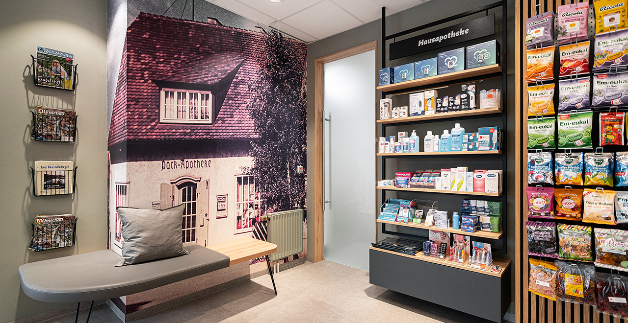 Regale in der Park-Apotheke, die Kosmetikprodukte und Nahrungsergänzungsmittel ausstellen. Die Regale sind gut organisiert, mit Beschilderungen, die auf verschiedene Produktkategorien hinweisen, wie „Kosmetik“ und „Nahrungsergänzung“. In der Mitte des Raums steht ein kleiner Tisch.