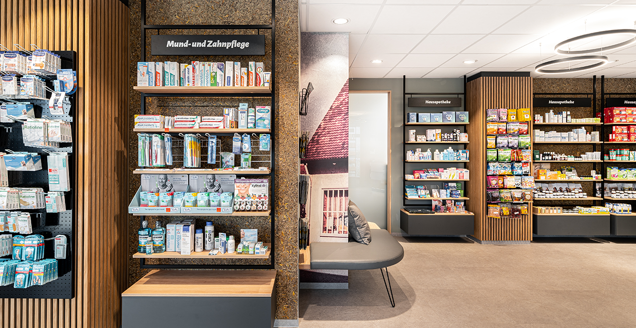 Blick auf einen Bereich der Park-Apotheke, in dem Regale für „Hausapotheke“ und „Mund- und Zahnpflege“ Produkte ausstellen. Auf der linken Seite befindet sich ein Regal mit Zahnpflegeprodukten und auf der rechten Seite Produkte für die Hausapotheke. In der Mitte des Raums steht eine Bank mit Kissen, die als Sitzgelegenheit dient.