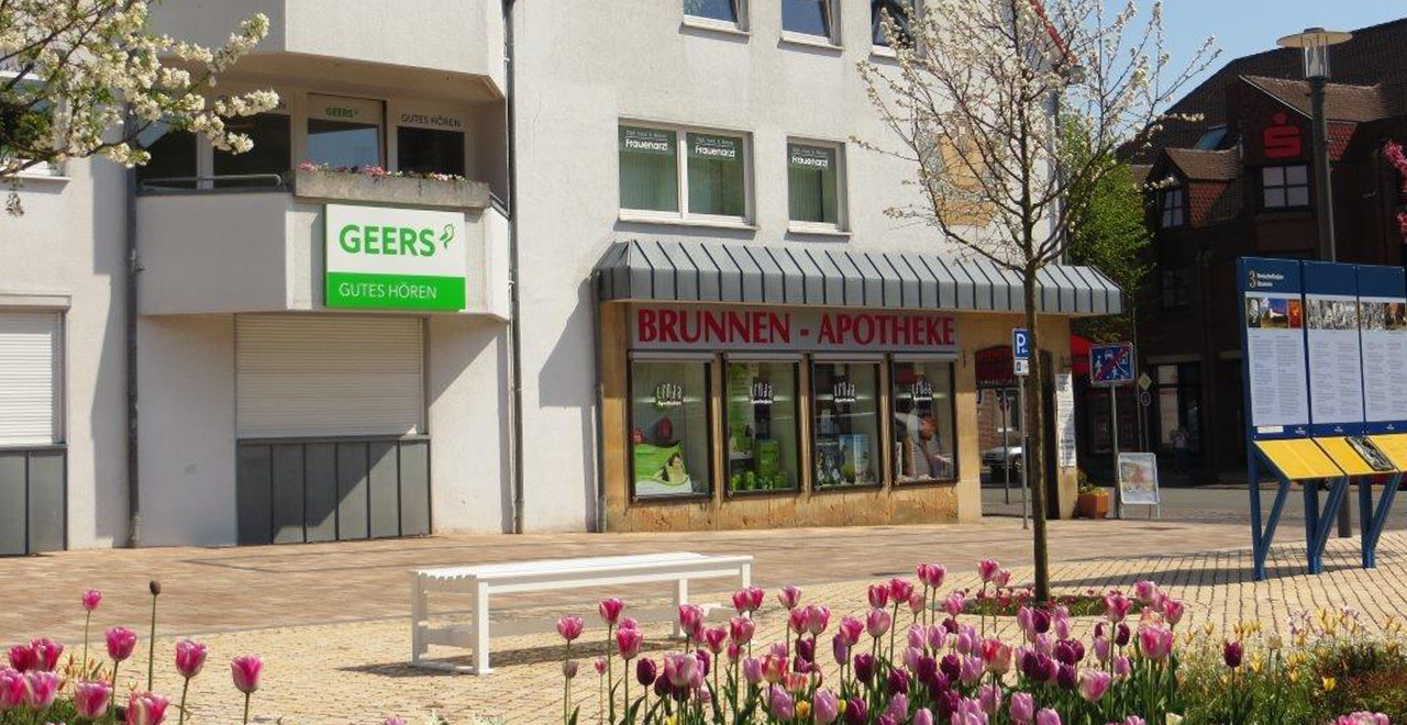 Außenansicht der Brunnen-Apotheke. Ein modernes Apotheken-Geschäft mit einem grünen Geers-Schild für Hörgeräte und einer ansprechenden Blumenpflanzung vor dem Gebäude. Die Fenster zeigen die Apotheke und ihren Servicebereich.