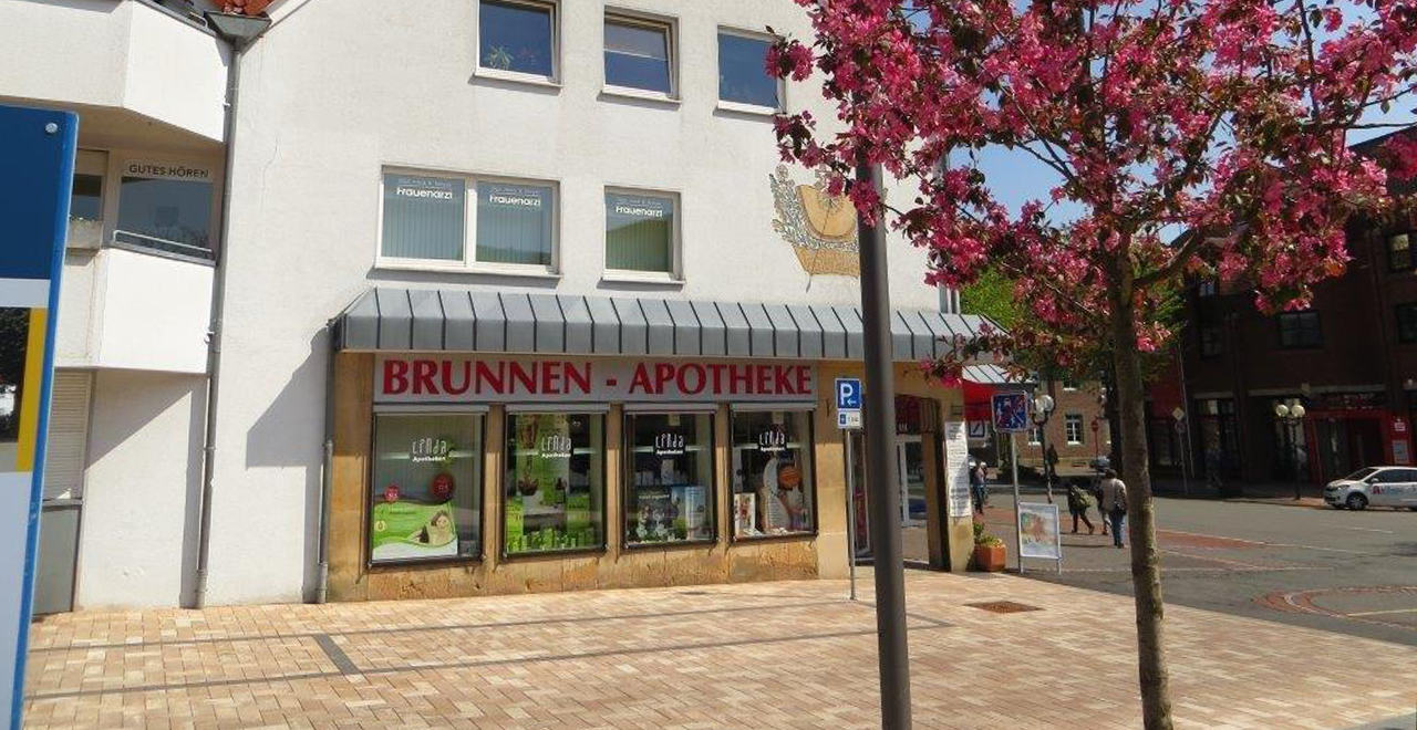 Blick auf die Brunnen-Apotheke mit einem roten Apotheken-Schild. Die Fenster sind mit Werbung und Informationsmaterial versehen. Eine rosa Blütenpflanze befindet sich auf der rechten Seite des Bildes.