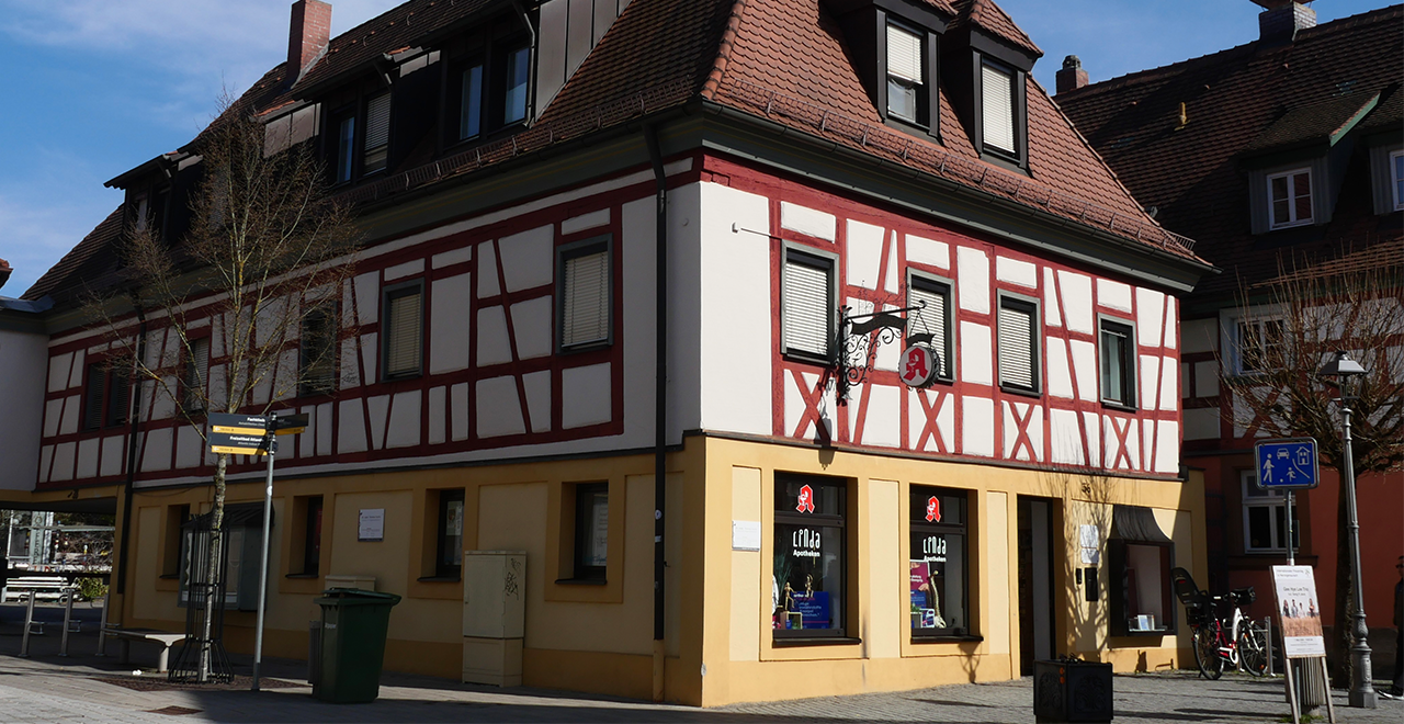 Ein zweistöckiges Gebäude mit weißer Fassade und roten Fachwerkbalken. Im Erdgeschoss befindet sich eine Apotheke mit Schaufenstern und dem typischen Apotheken-Symbol. Vor dem Haus stehen ein Fahrrad und Straßenschilder, daneben verläuft eine ruhige Straße.