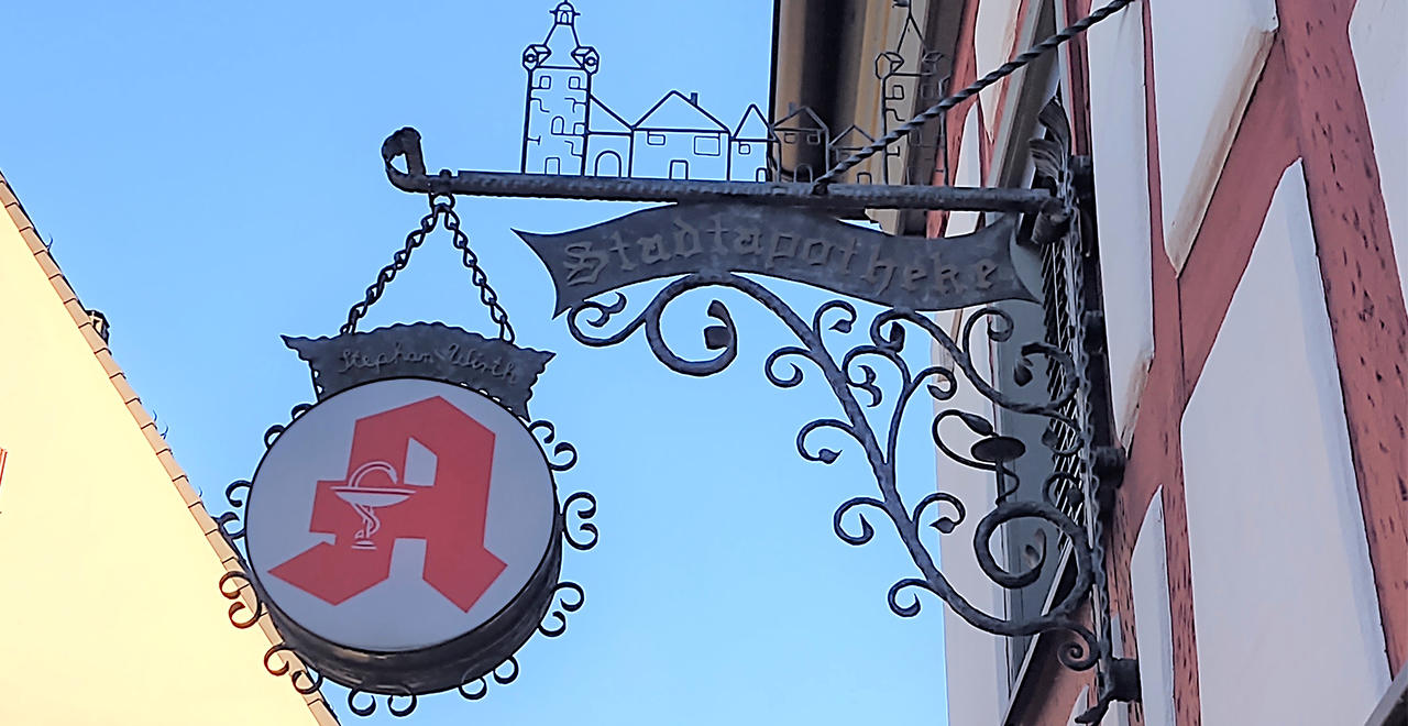 Außenansicht eines kunstvoll geschmiedeten Apothekenschilds mit rotem Apotheken A vor blauem Himmel; darüber der Schriftzug Stadtapotheke.