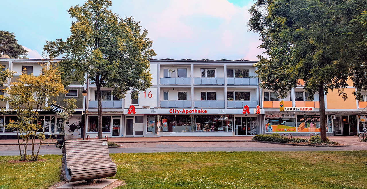 Ein modernes Gebäude mit weißen Fassaden und einem großen, gut sichtbaren Apothekenzeichen. Im Vordergrund sieht man einen kleinen Park mit Bäumen.