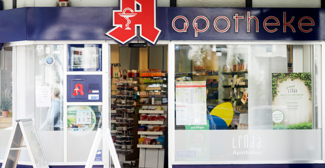 Frontansicht einer Apotheke mit dem Schriftzug 'apotheke' auf blauem Hintergrund. Ein großes rotes Apotheken-A hängt über dem Eingang. Durch die Tür und das Fenster sind Regale mit Medikamenten sichtbar. Im unteren Fensterbereich ist das Logo 'Linda Apotheken' zu sehen.
