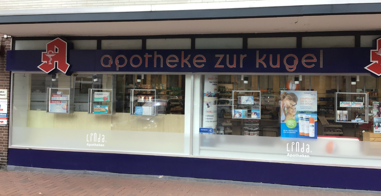 Breite Schaufensterfront der 'apotheke zur kugel'. Der Schriftzug ist in Kleinbuchstaben auf einem blauen Schild zu lesen. Links und rechts ist jeweils das rote Apotheken-A zu sehen. In den Schaufenstern sind verschiedene Werbeplakate und Medikamente ausgestellt. Unten steht 'Linda Apotheken'.
