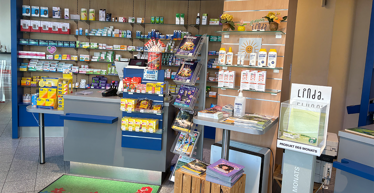 Innenansicht einer Apotheke mit einem Verkaufs- und Beratungstresen im Vordergrund. Hinter dem Tresen befinden sich Regale mit Medikamenten, Pflastern und Gesundheitsprodukten. Auf dem Tresen stehen kleine Verkaufsdisplays, Zeitschriften und Desinfektionsmittel. An der Wand sind Produkte und Informationsmaterialien ausgestellt.