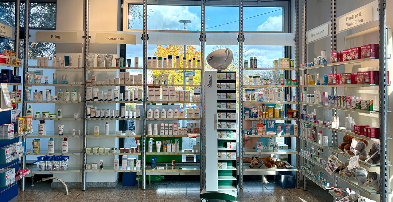 Regalwand mit großen Fenstern im Hintergrund, durch die Tageslicht hereinfällt. In den Regalen stehen Pflegeprodukte, Kosmetika, Vitamine und Artikel für Familien und Wohlbefinden. Die Bereiche sind mit Schildern wie „Pflege“, „Kosmetik“ und „Familien & Wohlfühlen“ gekennzeichnet. Einige Kuscheltiere sind im unteren Regal sichtbar, was eine freundliche Note vermittelt.