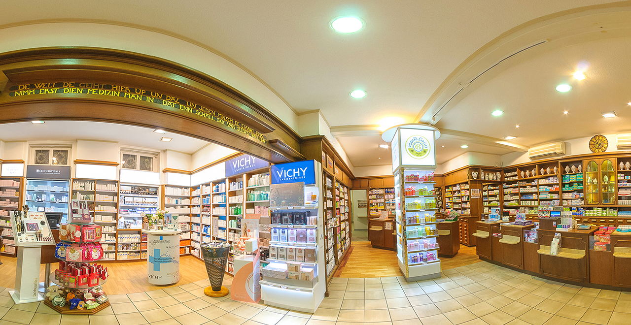 Innenansicht einer großzügig gestalteten Apotheke mit heller Beleuchtung und klassischer Holzverkleidung. Regale mit Pflegeprodukten, Kosmetika und Medikamenten säumen die Wände. Im Zentrum stehen Verkaufsständer bekannter Marken wie Vichy und Roger & Gallet.
