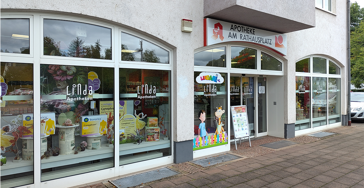 Außenansicht der Apotheke. In diesem Bild ist die Apotheke von außen mit einer klaren Sicht auf die Fensterfront zu sehen. Das Schaufenster ist dekoriert, und verschiedene Produkte sind sichtbar. Der Eingang ist deutlich erkennbar, und es gibt einen Werbeständer vor der Tür.