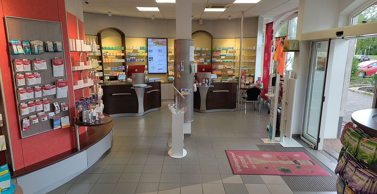Innenansicht der Apotheke. Der Raum zeigt Regale mit einer Vielzahl von Medikamenten und Gesundheitsprodukten. In der Mitte befindet sich der Empfangstresen mit digitalen Anzeigen und Computern. Der Raum ist gut strukturiert und die Regale sind sauber und ordentlich angeordnet.