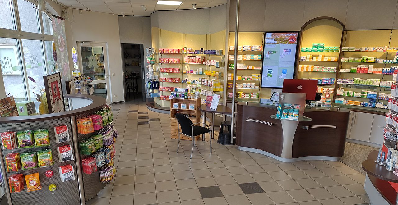 Innenansicht der Apotheke. Auf der linken Seite sind Regale mit verschiedenen Produkten wie Tees und Nahrungsergänzungsmitteln zu sehen. In der Mitte des Raumes befindet sich ein Empfangstresen mit einem Computer. Im Hintergrund sind weitere Regale mit Arzneimitteln sichtbar. Der Raum ist gut beleuchtet und modern gestaltet.