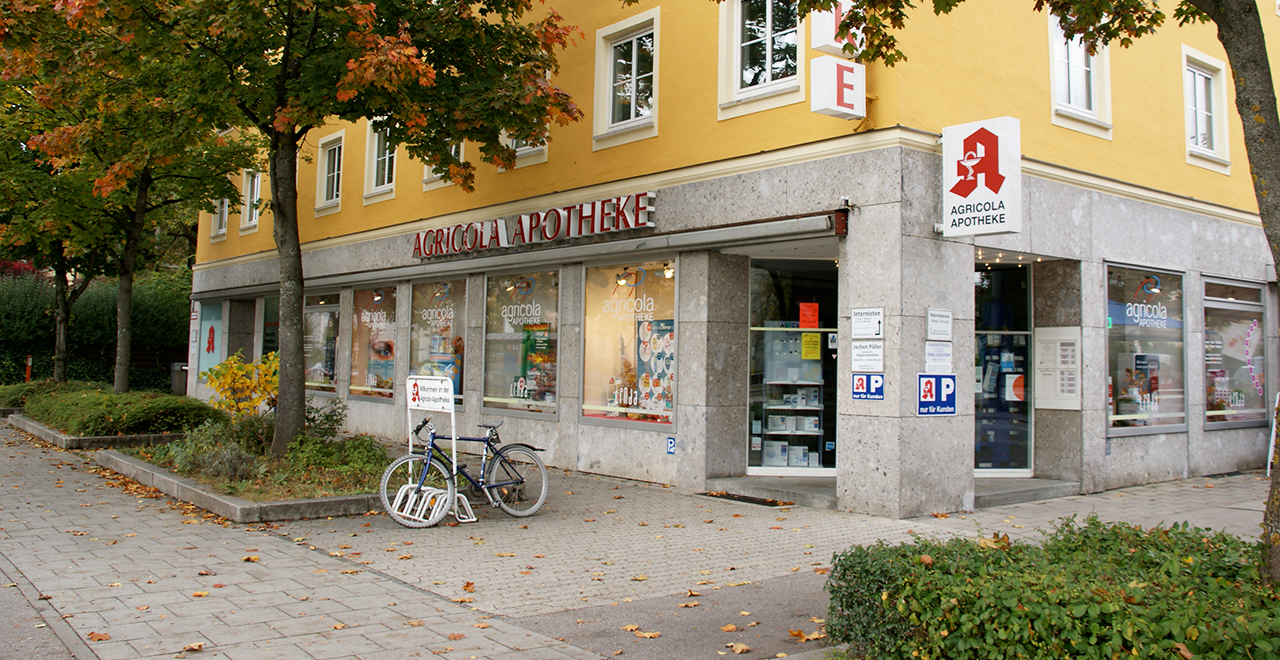 Außenansicht der Agricola Apotheke an einer Straßenecke. Das Gebäude hat eine gelbe Fassade mit grauen Steinelementen. Große Schaufenster zeigen Apothekenwerbung. Vor dem Eingang stehen zwei Fahrräder in einem Fahrradständer. Bäume mit herbstlich gefärbtem Laub säumen den Gehweg.