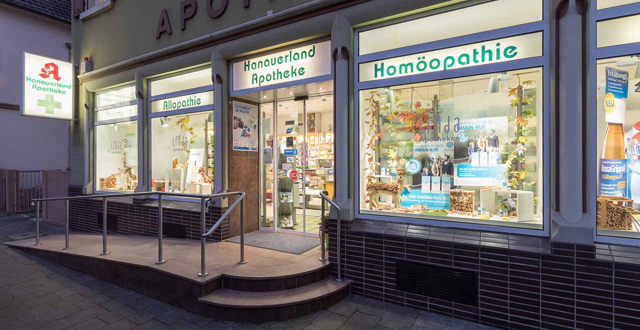 Außenansicht der Hanauerland Apotheke bei Nacht. Das Geschäft hat zwei Schaufenster mit der Aufschrift 'Allopathie' und 'Homöopathie'. Das Logo der Apotheke ist gut sichtbar, und die Beleuchtung hebt die Schaufenster hervor.