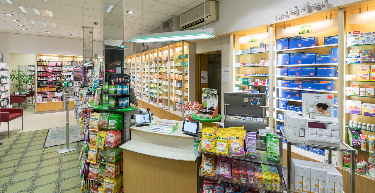Nahaufnahme des Kassenbereichs in der Hanauerland Apotheke. Auf dem Tresen sind diverse Produkte und Verpackungen ausgestellt. Im Hintergrund sind weitere Regale mit Medikamenten und Pflegeprodukten zu sehen.