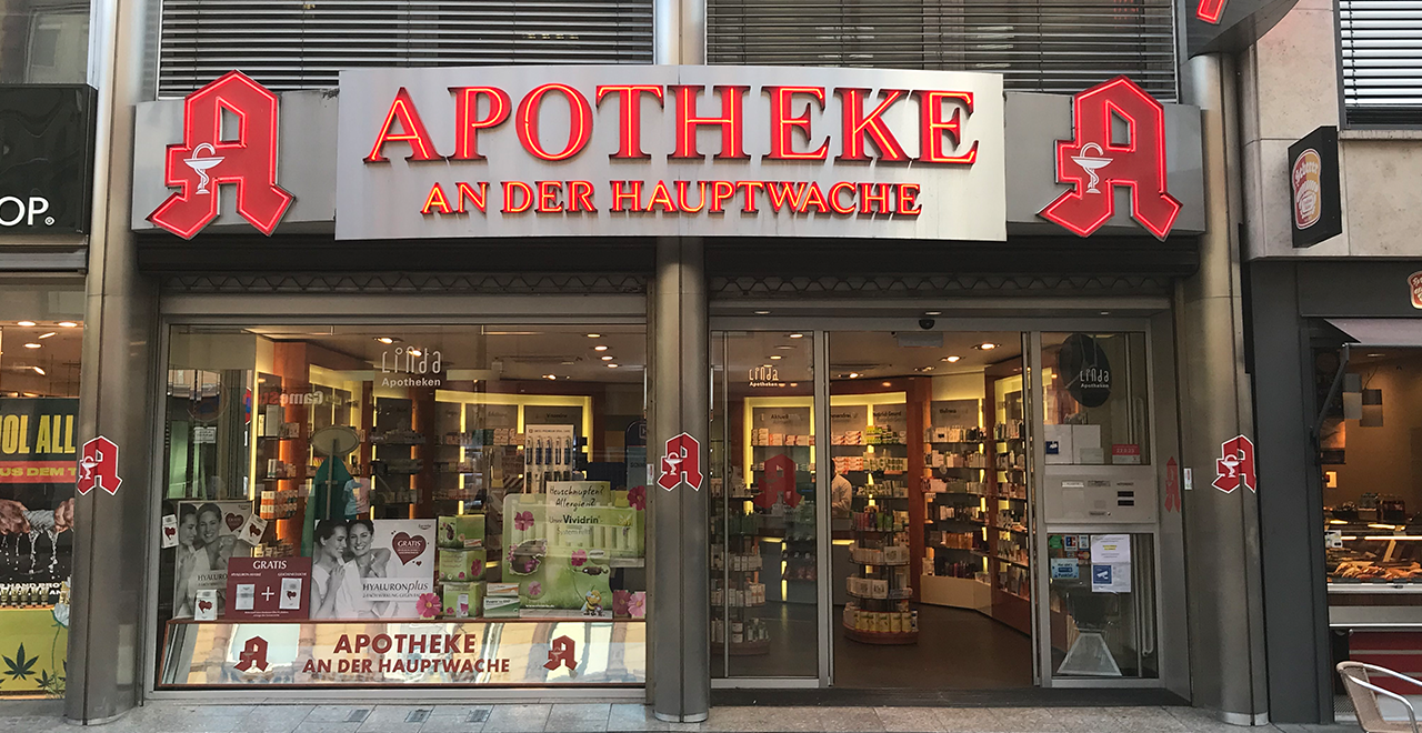 Außenansicht einer Apotheke mit dem Schriftzug ‚APOTHEKE AN DER HAUPTWACHE‘ in großen roten Leuchtbuchstaben. Die Fassade ist modern, mit großen Glasfenstern, durch die man Regale und Produkte im Inneren sehen kann. Auf beiden Seiten und in der Mitte der Eingangstür sind rote Apotheken-Logos angebracht.