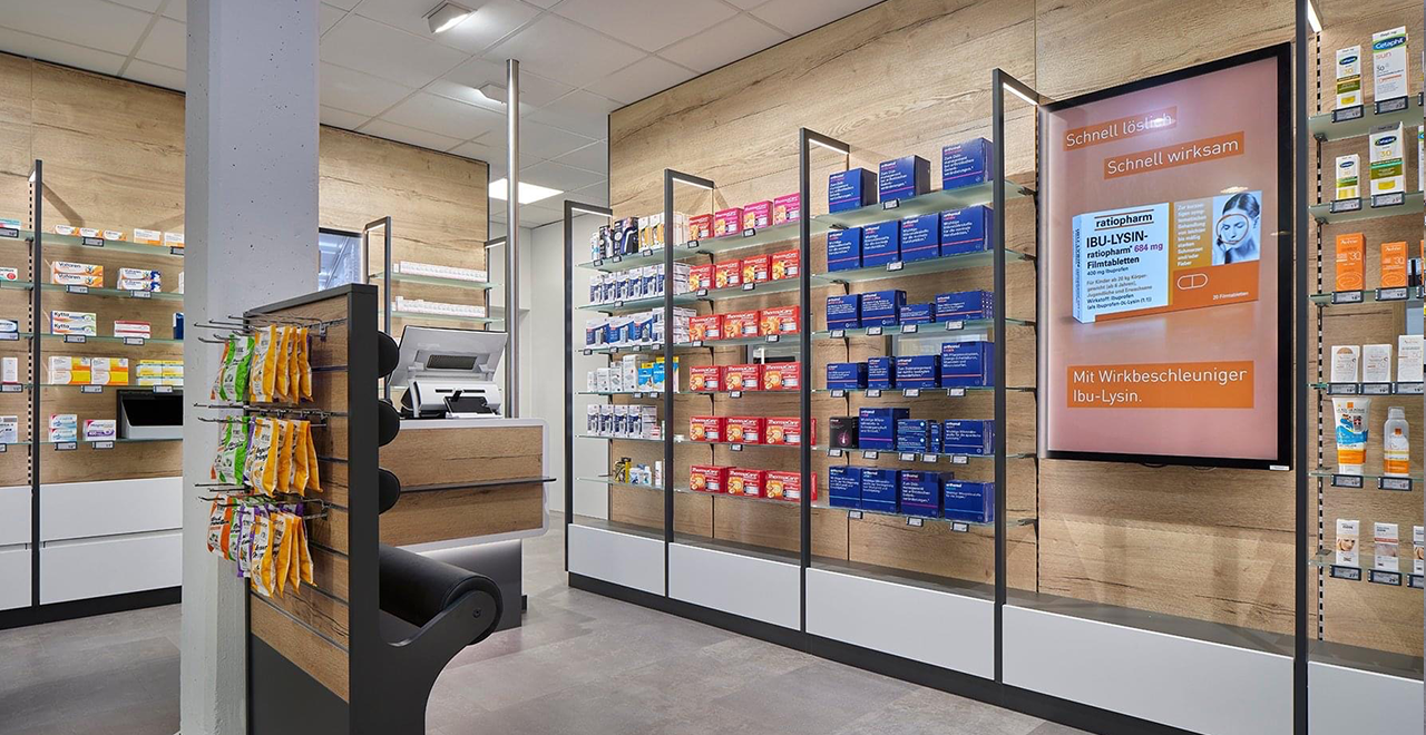 Beratungstheke in einer Apotheke mit beleuchtetem Medikamentenregal und Werbebildschirm.