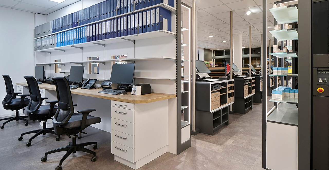 Das Bild zeigt den Bürobereich einer Apotheke mit mehreren nebeneinander aufgereihten Schreibtischplätzen. Jeder Arbeitsplatz ist mit einem Bildschirm, Telefon und Büroausstattung ausgestattet. Über den Tischen befinden sich weiße Regalbretter mit zahlreichen blauen und grauen Aktenordnern. Rechts im Bild sind der Verkaufsraum der Apotheke sowie ein automatisiertes Lagersystem mit Medikamenten sichtbar.