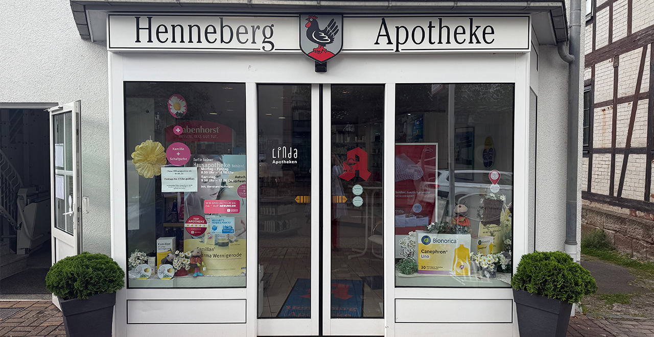 Frontansicht der Henneberg Apotheke mit einer großen Glastür, weißen Wänden und einem Schild mit einem Hahn-Logo. Im Fenster sind Anzeigen für verschiedene Gesundheitsprodukte zu sehen, die Tür wird von dekorativen Pflanzen flankiert.