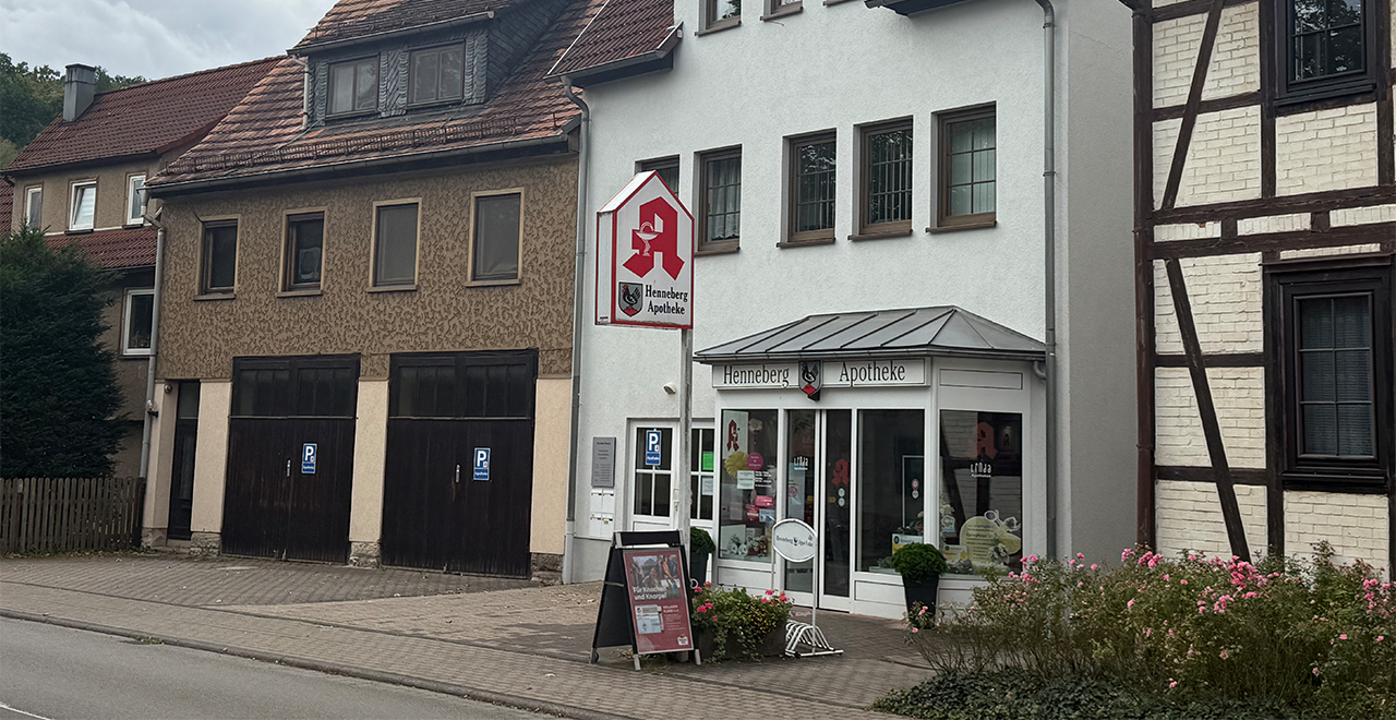 Ein weiterer Blick auf die Henneberg Apotheke von der Straße aus, mit dem weißen Gebäude und dem roten Hahn-Schild über dem Eingang. In der Nähe befinden sich Parkplätze und einige grüne Pflanzen vor dem Geschäft.