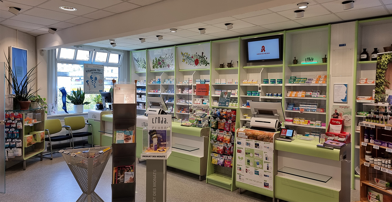 Das Bild zeigt eine moderne Apotheke. In der Mitte befinden sich zwei große Computerarbeitsplätze mit Kassen, umgeben von Regalen mit verschiedenen Medikamenten, Nahrungsergänzungsmitteln und Gesundheitsprodukten. Über den Regalen sind Pflanzenmuster abgebildet. Durch die Fenster im Hintergrund ist viel natürliches Licht zu sehen. Es gibt Sitzmöglichkeiten und Informationsmaterialien zur Verfügung, die die Kunden bei ihrem Besuch unterstützen. Ein Bildschirm an der Wand zeigt Werbung und Informationen. Die Einrichtung ist hell und freundlich gestaltet.