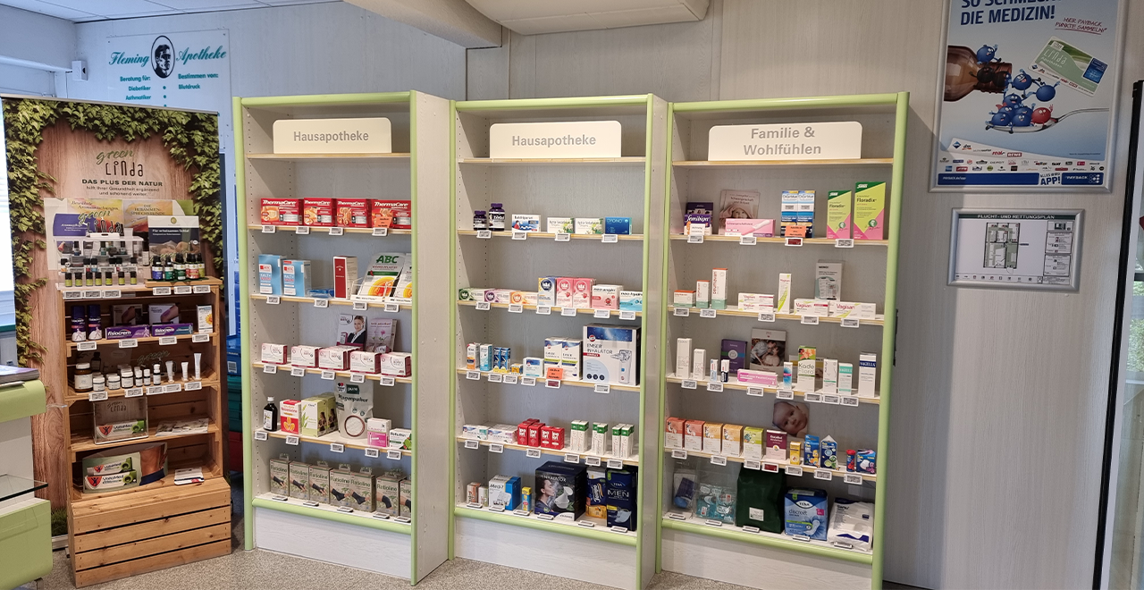 Auf diesem Bild sieht man einen weiteren Teil der Apotheke. Es gibt drei Regale, die mit verschiedenen Produkten für die Hausapotheke sowie für Familie und Wohlbefinden gefüllt sind. Das Design ist schlicht und übersichtlich, mit einer klaren Beschriftung der Produktkategorien. Links ist ein Holzregal mit Naturprodukten und Heilmitteln zu sehen. An der Wand hängt ein Poster, das auf eine App für die Apotheke hinweist. Die Regale sind hell beleuchtet und ordentlich strukturiert.