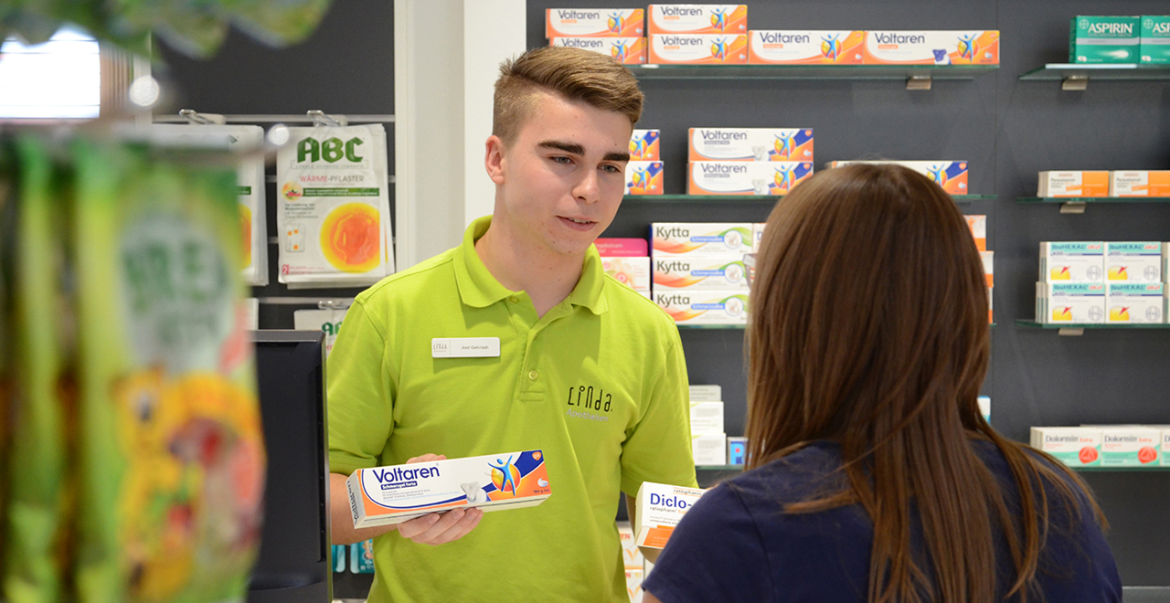 Apothekenmitarbeiter berät eine Kundin und zeigt eine Packung Voltaren Schmerzgel.