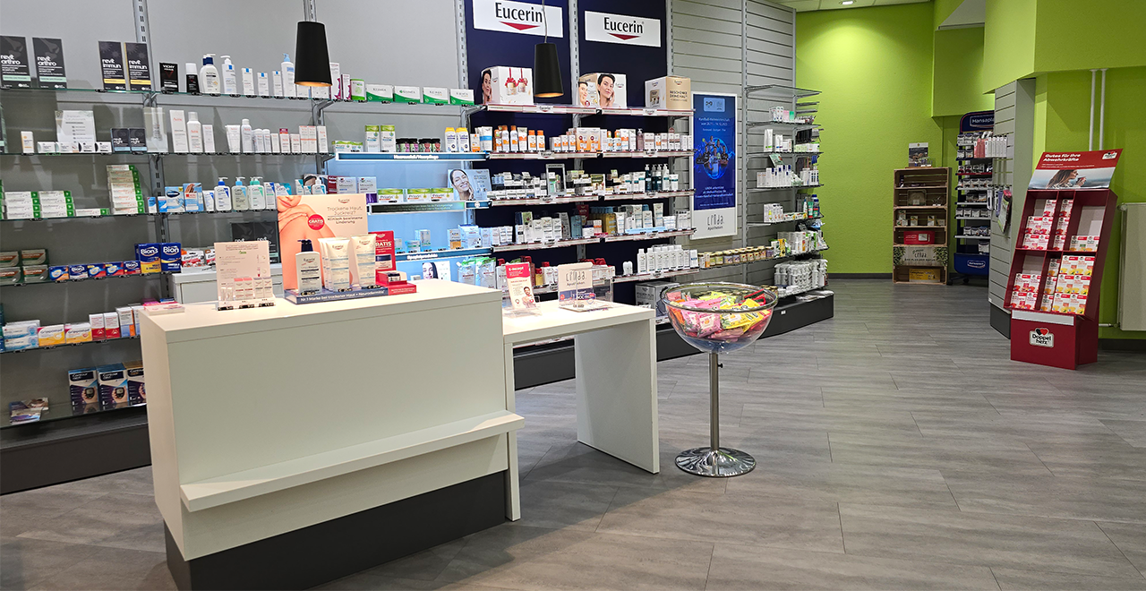 Apothekenbereich mit Fokus auf Hautpflegeprodukte. Im Vordergrund steht ein weißer Beratungstisch, dahinter Glasregale mit Cremes und Lotionen von Marken wie Eucerin. Rechts steht eine große transparente Schale mit bunt verpackten kleineren Produkten. Links und rechts ziehen sich Regale mit Medikamenten die Wand entlang. Der Laden ist hell, modern und gut zugänglich gestaltet.