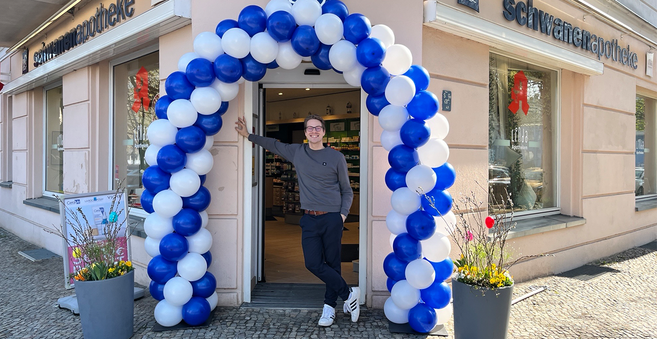 Der Inhaber steht lächelnd im Eingang der Schwanen Apotheke und lehnt sich mit einer Hand an den Türrahmen. Über ihm spannt sich ein großer Bogen aus blauen und weißen Luftballons. Links und rechts neben dem Eingang stehen Pflanzkübel mit Blumen. Durch die offene Tür ist der Innenraum der Apotheke mit Regalen erkennbar.